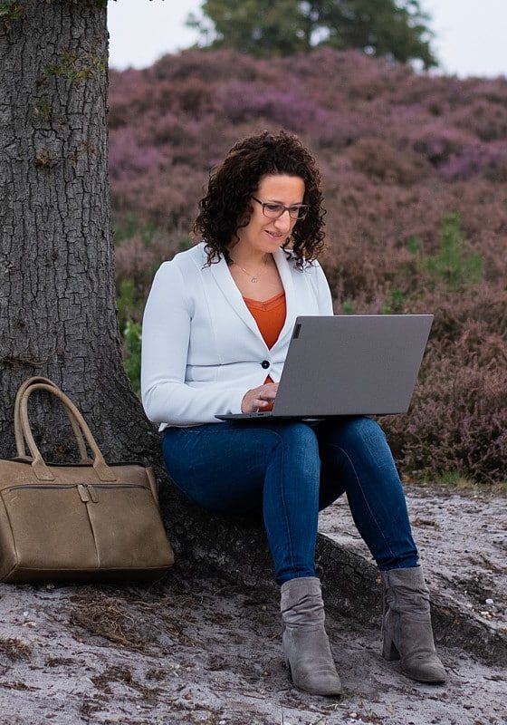 Femke, werkend op een laptop tegen een boom aan, in een heideveld.